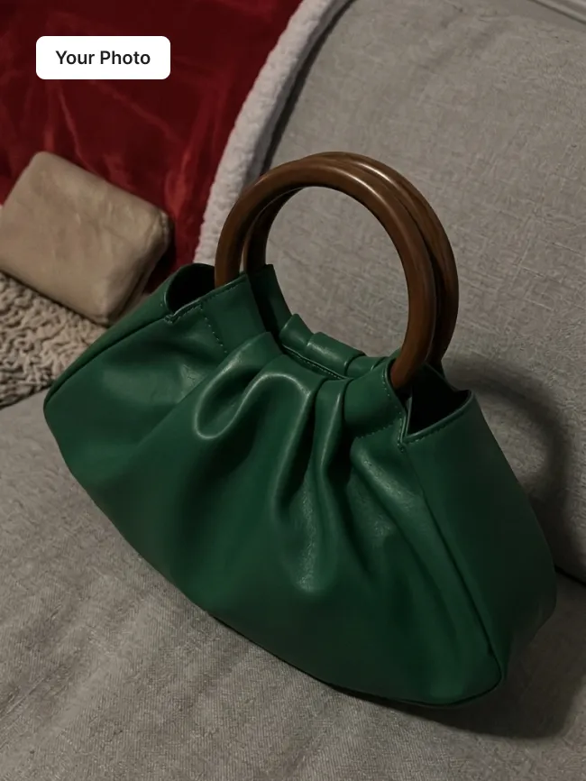 Plain phone photo of a green pleated handbag with a wooden top handle on a grey seat