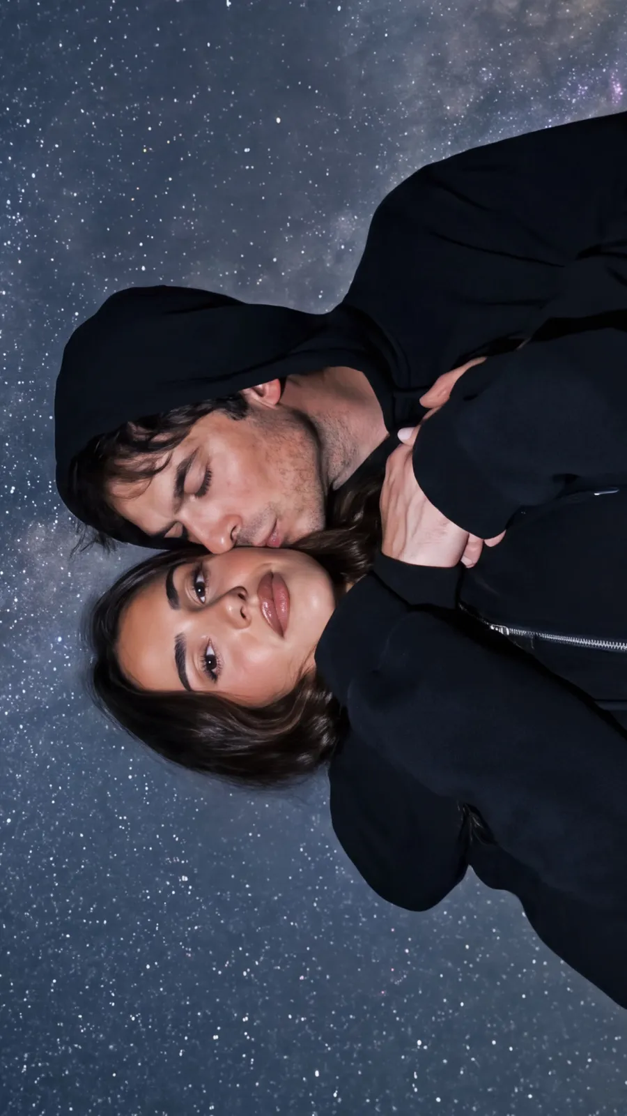 Couple in black hoodies lying head to head under the Milky Way, shot from directly above